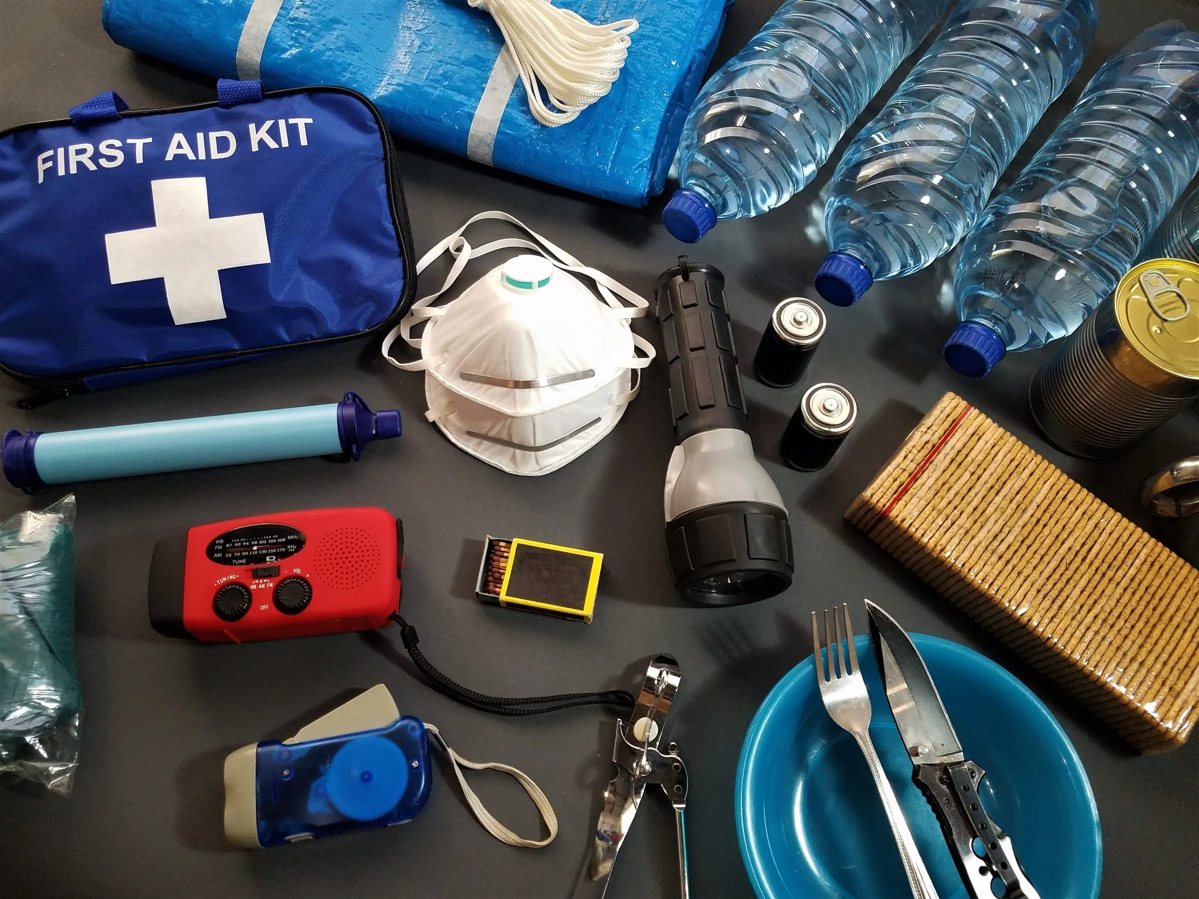 A first aid kit laid out with emergency supplies: bottled water, a flashlight, a whistle, a hand-crank radio, and more